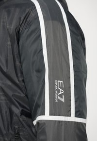 Manseta jachetei negre Emporio Armani EA7 cu dungi reflectorizante albe și logo, evidențiind țesătura texturată și detaliile fermoarului.