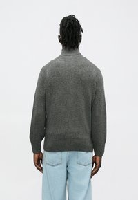 Sivý pletený sveter s vysokým golierom, rebrovanými manžetami a lemom. Má textúrovaný povrch a voľný strih. Nosí sa so svetlomodrými džínsami.