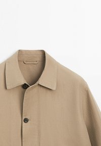 Beige jakke med krave, synlig øverste knap og enkel syning mod en hvid baggrund, der viser øvre front og kraveområde.