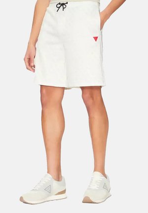 Personne portant un short blanc avec un cordon noir et des baskets blanches avec un logo triangulaire sur un fond uni.