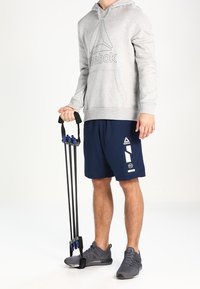 Šedá mikina s logem Reebok, tmavě modré šortky s bílými akcenty, držící černý a modrý odporový pás, sportovní obuv, neutrální pozadí.