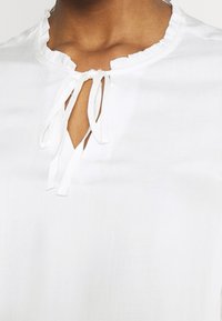 Blusa bianca con collo rotondo e volant, dotata di apertura a chiave, fissata da due laccetti frontali, realizzata in tessuto leggero.