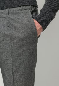 Graue Fischgrätenhose mit strukturierter Oberfläche, ausgestattet mit seitlichen Taschen und taillierter Passform, kombiniert mit einem dunkel gestrickten Pullover.