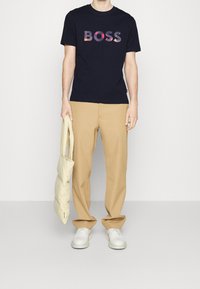 Marinblå bomulls-T-shirt med "BOSS"-logotyp i färgglad tryck, kombinerad med beiga byxor och vita skor, håller en ljusbeige puffer-väska.