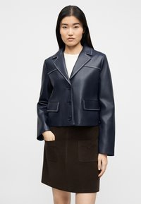 Femme portant une veste en cuir bleu marine avec des coutures blanches et une jupe marron foncé avec des poches avant, sur un fond uni.