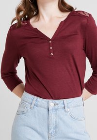 Burgundy långärmad topp med V-ringning, treknappars placket och spetsdetaljer på axlarna. Matchad med ljusblå denimshorts med hög midja.