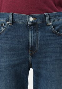 Acercamiento a unos vaqueros de mezclilla azul con un botón plateado y costuras naranjas, llevados con una camisa color burdeos metida por dentro.