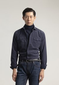Camisa de botón azul marino con dos bolsillos en el pecho, combinada con un cuello alto oscuro y jeans azules, con una tela texturizada y un diseño ajustado.