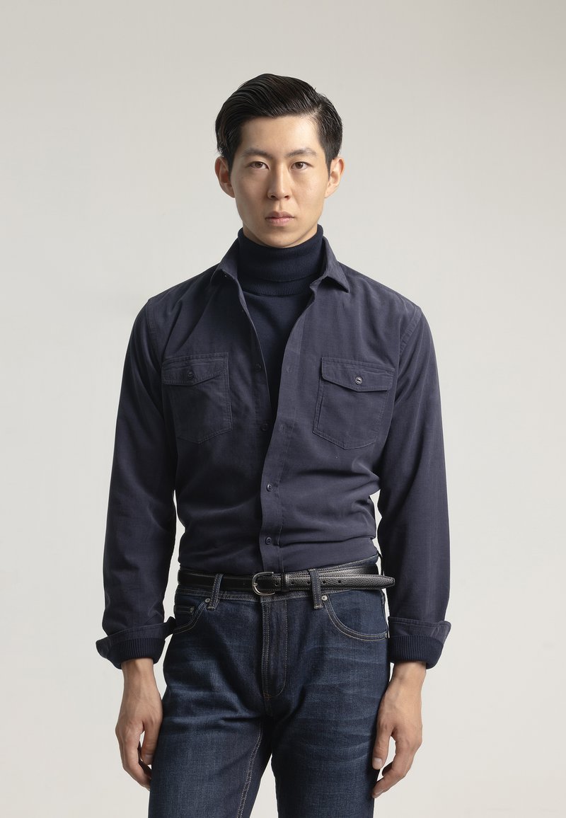 Camisa de botón azul marino con dos bolsillos en el pecho, combinada con un cuello alto oscuro y jeans azules, con una tela texturizada y un diseño ajustado.