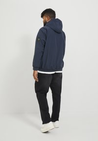 Chaqueta con capucha de color navy con textura suave y puños elásticos, combinada con pantalones negros y zapatillas blancas; parche con el logotipo en la manga.