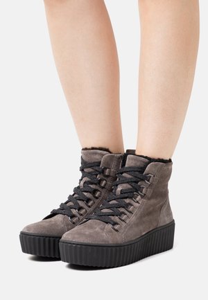 Szare zamszowe botki na platformie do kostki z czarnymi grubymi sznurowadłami, czarnymi żłobionymi podeszwami i miękką wyściółką noszone na nagich nogach.