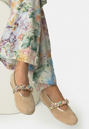 Chaussures plates en daim beige avec des brides ornées de pierres précieuses multicolores, portées avec un pantalon large à motifs floraux.