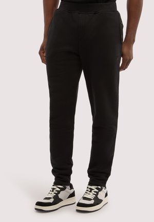 Person iført sorte tapered sweatpants med sidelommer og sort-hvide sneakers, stående mod en ensfarvet baggrund.