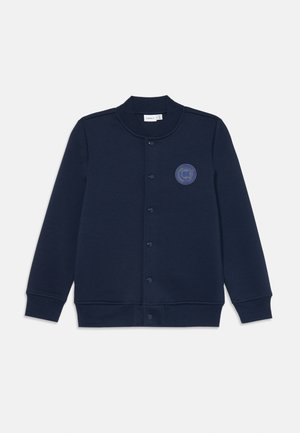 Veste sweat bleu marine avec col rond, boutons pression sur le devant, poignets côtelés et détail de logo circulaire sur la poitrine gauche.