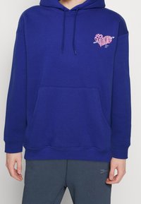 Modrá fleecová mikina s kapucí, přední kapsou, stahovací šňůrkou na kapuci a růžovým srdíčkem na levé straně hrudi. Žebrované lemy na rukávech a spodním okraji.