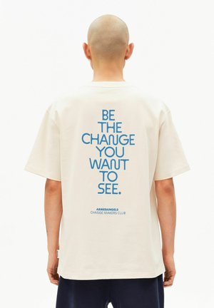 Cream-colored t-shirt with a loose fit, featuring blue text on the back: "BE THE CHANGE YOU WANT TO SEE." Brand logo above text.