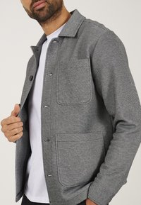 Grijze, gestructureerde jas met een kraaghalslijn, twee voorzakken en knoopsluitingen, gedragen over een wit shirt.