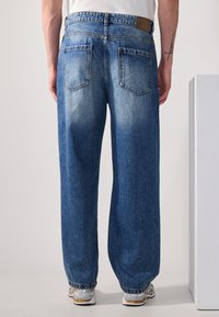 Jeans de denim azul deslavado de pernera ancha con textura lavada, diseño de cinco bolsillos y puños elásticos. La parte trasera presenta la etiqueta de la marca y detalles de costura.