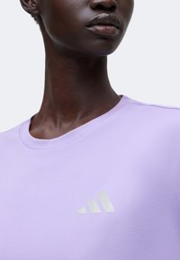 Gros plan d'une personne portant un t-shirt Adidas violet clair avec le logo visible sur la poitrine, montrant le cou et le bas du visage.