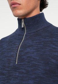 Navy blå zip-up genser med høy krage og strukturstrikk. Har en sølvfarget metallglidelås som dekorasjon foran.