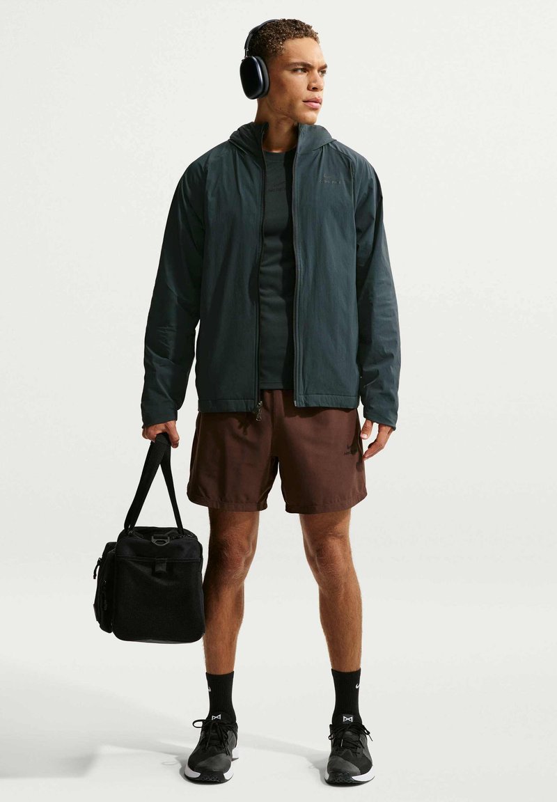 Chaqueta deportiva verde oscuro, pantalones cortos deportivos marrones, zapatillas negras con logo, llevando una bolsa negra, usando auriculares negros.