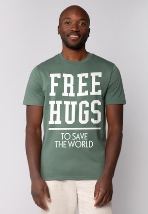 Grünes Baumwoll-T-Shirt mit auffälligem weißen Text: "KOSTENLOSE UMARMUNGEN" und "UM DIE WELT ZU RETTEN." Kurzarm- und Rundhalsausschnitt-Design.