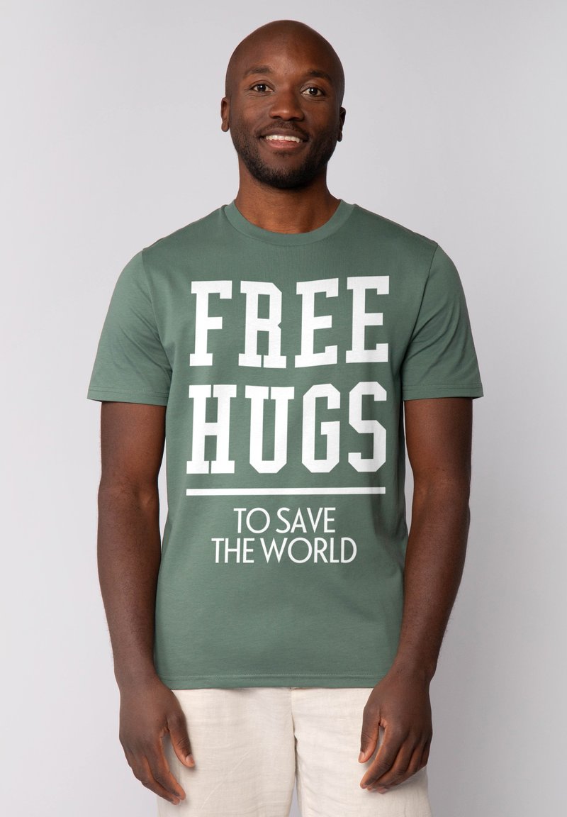Groene katoenen T-shirt met opvallende witte tekst: "GRATIS KNUFFELS" en "OM DE WERELD TE REDDEN." Korte mouwen en een ronde hals.