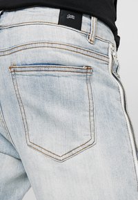 Hellblaue Jeans aus Denim verfügen über einen Reißverschluss an der Seite, hintere Taschen mit kontrastierendem Stich und ein schwarzes Markenpatch oberhalb des Bundes.