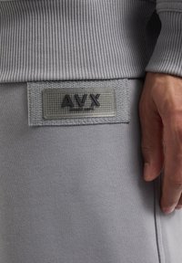 Grijs sweatshirtmateriaal met geribbelde manchetten en een textuur patch met het logo "AVX" op een gladde achtergrond. Close-up van het materiaal en de details.