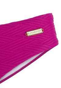 Magenta simklädestyg med ett texturerat zigzagmönster, prytt med en guldfärgad "sunseeker"-tagg för varumärkesidentitet. Mjuk innerfoder.