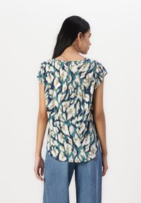 Blouse à manches courtes en teal avec un motif abstrait crème et marine, dotée d'une fermeture éclair au dos et d'un ourlet incurvé. Finition en tissu doux.
