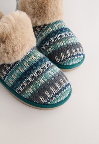 Chaussons tricotés présentant un motif multicolore dans des tons de vert, bleu et gris, avec un revers en fausse fourrure et une semelle plate en caoutchouc.