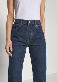 Jean en denim bleu taille haute avec une coupe classique, comportant cinq poches, des détails en cuivre et des coutures subtiles.