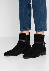 Stivali alla caviglia in suede nero con cerniera laterale e fascia decorativa in pelle, indossati con jeans in denim blu tagliati su un pavimento bianco liscia.