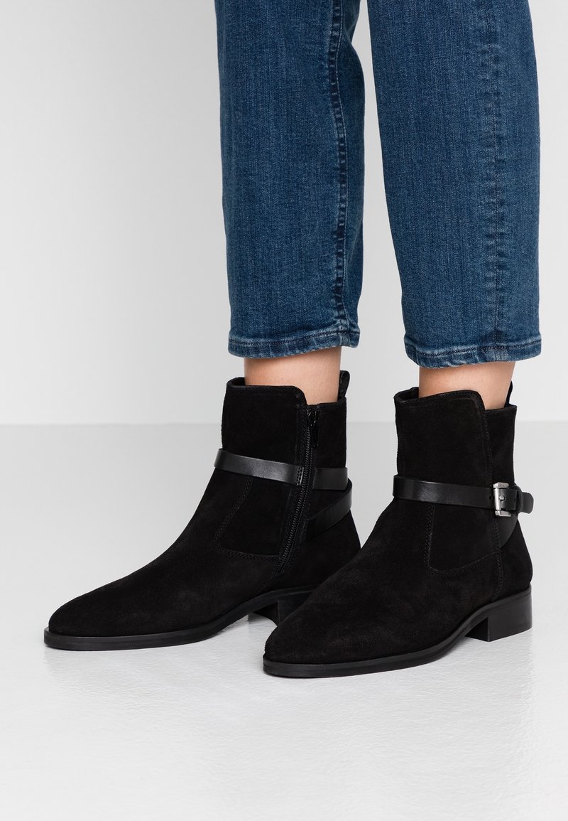 Stivali alla caviglia in suede nero con cerniera laterale e fascia decorativa in pelle, indossati con jeans in denim blu tagliati su un pavimento bianco liscia.
