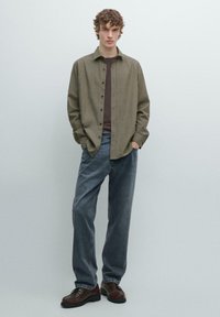 Koszula na guziki z oliwkowej tkaniny, zestawiona z ciemnobrązowym t-shirtem i luźnymi szarymi dżinsami, uzupełniona brązowymi butami.