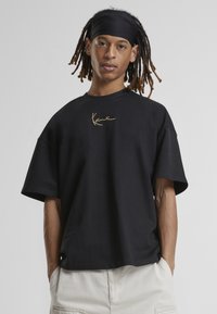 Černé oversized tričko vyrobené z bavlny, s kulatým výstřihem a krátkými rukávy. Zlatý podpisový detail na oblasti hrudníku.