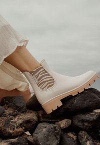 Botín blanco a los tobillos con un acabado suave, suela beige y laterales elásticos con estampado animal. Posicionado sobre rocas negras, parcialmente cubierto por una tela ligera.