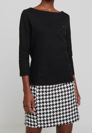 Mujer vestida con una blusa negra de manga larga y una falda de patrón pata de gallo en blanco y negro, posando contra un fondo gris liso.