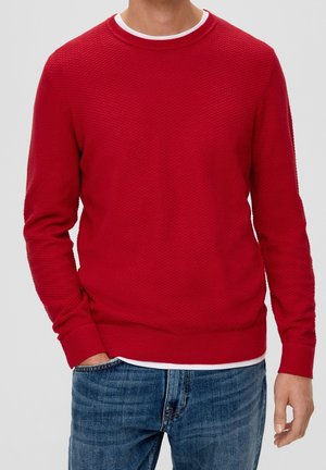 Mand iført en tekstureret rød langærmet sweater over en hvid skjorte, med blå jeans, stående mod en ensartet lys baggrund.