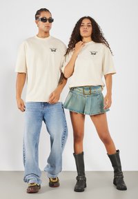 Bežová grafická trička, široké modré džíny, džínová plisovaná sukně a černé vysoké boty. Sluneční brýle nosí mužský model.