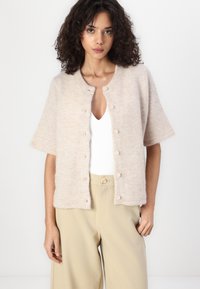 Cardigan corto beige a maniche corte realizzato in un materiale morbido e testurizzato. Presenta un collo rotondo e sei bottoni lungo la parte frontale.