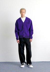 Cardigan violet cu gât în V și închidere cu nasture, dungi negre în partea de jos, purtat peste o cămașă albă și asortat cu pantaloni negri și adidași albi.