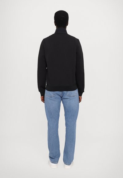 Svart hettegenser med høy krage og ribbede mansjetter, parret med lyseblå rett jeans. Bakfra vises en glatt tekstur.