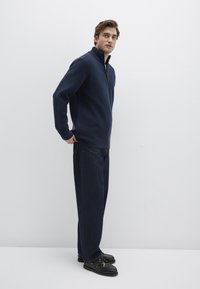 Navy fleece pullover with a zip collar, paired with loose denim trousers, and black shoes; smooth texture and casual design.