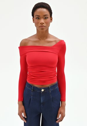 Donna con capelli corti che indossa un top rosso a maniche lunghe scoperte sulle spalle e jeans blu scuro a vita alta, in piedi davanti a uno sfondo bianco semplice.