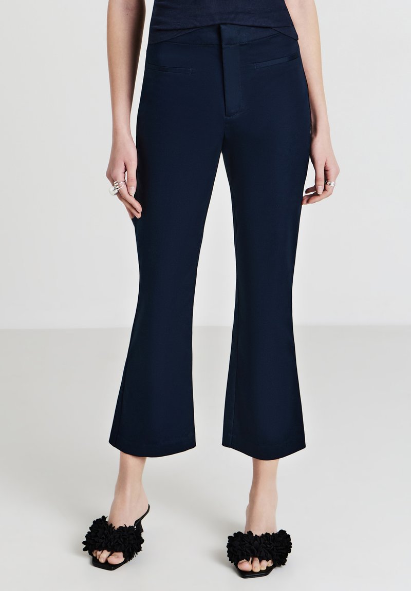 Pantaloni svasati blu navy con una texture liscia e tasche frontali, abbinati a sandali neri a scivolo decorati con fiori in tessuto.