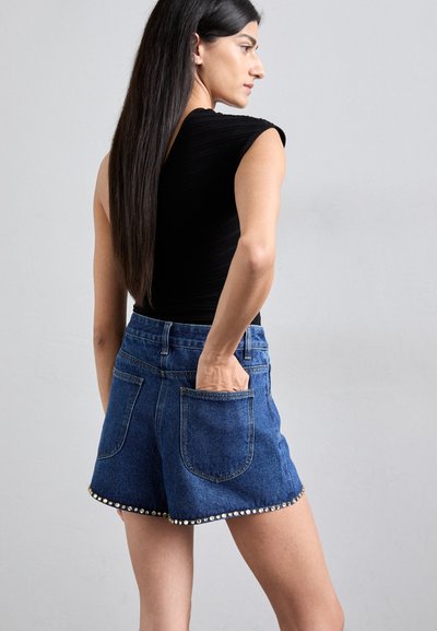 Pantaloni shorts in denim con vita alta, caratterizzati da un orlo ondulato decorato con borchie argento. Il tessuto è di un blu medio con una texture liscia.