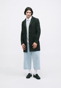 Černý kostkovaný kabát s klopovým límcem, šedá košile s knoflíky, světle modré volné džíny, bílé ponožky a černé slip-on boty.