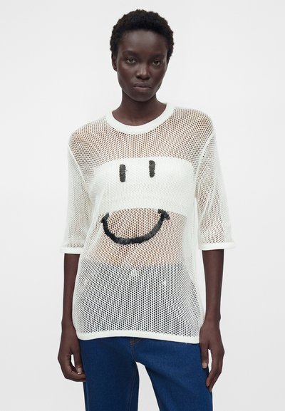 Άτομο με σκουρόχρωμο δέρμα που φοράει λευκό τοπ από δίχτυ με μαύρο σχέδιο χαμογελαστού προσώπου και μπλε τζιν, στέκεται μπροστά σε απλό φόντο.